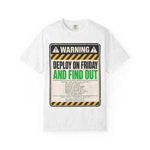 Pode incluir: Uma t-shirt branca de manga curta com um gráfico retangular que lembra um sinal de aviso. O design inclui o texto WARNING, DEPLOY ON FRIDAY AND FIND OUT e uma lista de efeitos secundários, emoldurado por faixas amarelas e pretas de perigo.