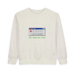 Op de afbeelding: Een lichtbeige sweatshirt met een afbeelding van een computerfoutmelding. De melding luidt "System Alert" met "404: Sleep Not Found" in een pop-upvenster. Daaronder staat de tekst "404: Sleep Not Found" in het groen gedrukt.