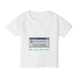 Könnte beinhalten: Weißes T-Shirt mit einem humorvollen Grafikdesign. Das Design zeigt eine Computerfehlermeldung mit der Aufschrift "404: Sleep Not Found". Das Design enthält ein blaues Fenster mit dem Text "System Alert" und den Optionen "OK" und "Try Again".