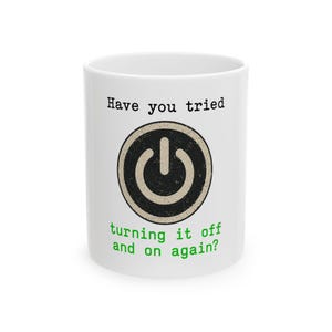 Puede incluir: Taza de cerámica blanca con el texto "Have you tried turning it off and on again?" en negro y verde. La taza presenta un gráfico de botón de encendido negro y beige.