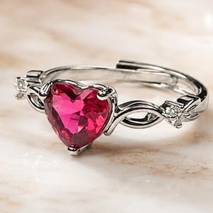 Könnte beinhalten: Ein silberfarbener Ring mit einem markanten herzförmigen roten Edelstein in der Mitte. Die Ringschiene hat ein offenes, verstellbares Design mit filigranen Metallarbeiten und kleinen klaren Steinen, die beidseitig am Herz eingefasst sind.