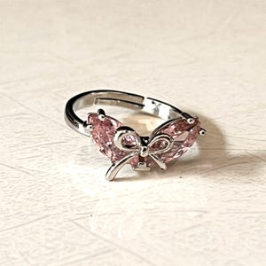 May include: A silver-toned adjustable ring featuring a central metal bow design flanked by two pear-shaped, light pink faceted stones. The band is open at the back, allowing for size adjustment, and the metal has a polished, reflective finish.