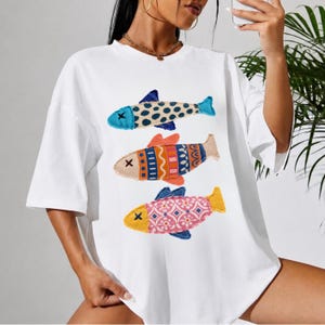 Peut inclure: Un t-shirt blanc à manches courtes orné d'un imprimé graphique représentant trois poissons stylisés empilés verticalement. Le poisson du haut est bleu à pois blancs, celui du milieu présente des motifs géométriques orange et bleus, et le bas est rose à motifs floraux.