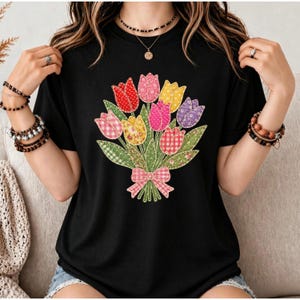 Könnte beinhalten: Ein schwarzes Kurzarm-T-Shirt mit einer zentralen Grafik eines Tulpenstraußes. Die Tulpen sind in verschiedenen Mustern wie Vichy-Karos und Blumenprints in Rot, Pink, Gelb und Violett dargestellt, ergänzt durch grüne Blätter und eine rosa Schleife.