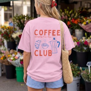 May include: A light pink t-shirt featuring the text COFFEE CLUB in orange, with blue line art illustrations of a croissant, a coffee cup, and a moka pot, along with the text everyday 7am-11am printed on the back of the garment.