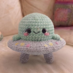 Può includere: Un giocattolo alieno all'uncinetto di colore verde chiaro con una base grigia a forma di UFO. L'alieno ha occhi neri, guance rosa e una bocca sorridente. La base dell'UFO ha pois colorati e gambe grigie.