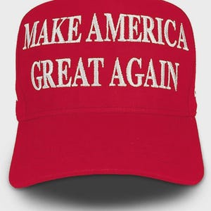 May include: A bright red baseball cap featuring the text MAKE AMERICA GREAT AGAIN embroidered in white capital letters across the front panel. The cap has a structured crown and a curved brim, with the stitching clearly visible on the fabric.