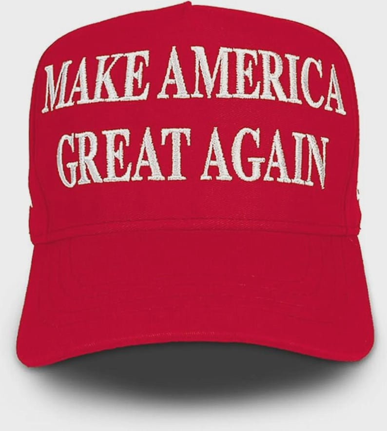 May include: A bright red baseball cap featuring the text MAKE AMERICA GREAT AGAIN embroidered in white capital letters across the front panel. The cap has a structured crown and a curved brim, with the stitching clearly visible on the fabric.