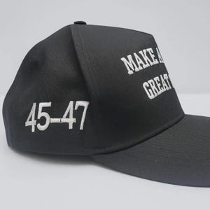 Op de afbeelding: Een zwarte honkbalpet met wit geborduurde tekst op het voorpaneel die MAKE A GREAT leest en de cijfers 45-47 in het wit geborduurd op het zijpaneel. De pet heeft een gestructureerde kroon en een gebogen klep gemaakt van zwarte stof.