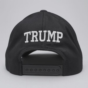 Op de afbeelding: Een zwarte baseballpet met het woord TRUMP in witte hoofdletters geborduurd op het achterpaneel. De pet is voorzien van een zwarte plastic snapback-sluiting aan de achterzijde, waardoor de maat verstelbaar is voor een goede pasvorm.