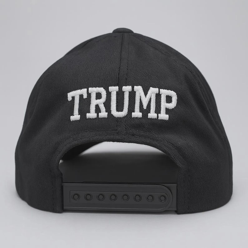 Op de afbeelding: Een zwarte baseballpet met het woord TRUMP in witte hoofdletters geborduurd op het achterpaneel. De pet is voorzien van een zwarte plastic snapback-sluiting aan de achterzijde, waardoor de maat verstelbaar is voor een goede pasvorm.