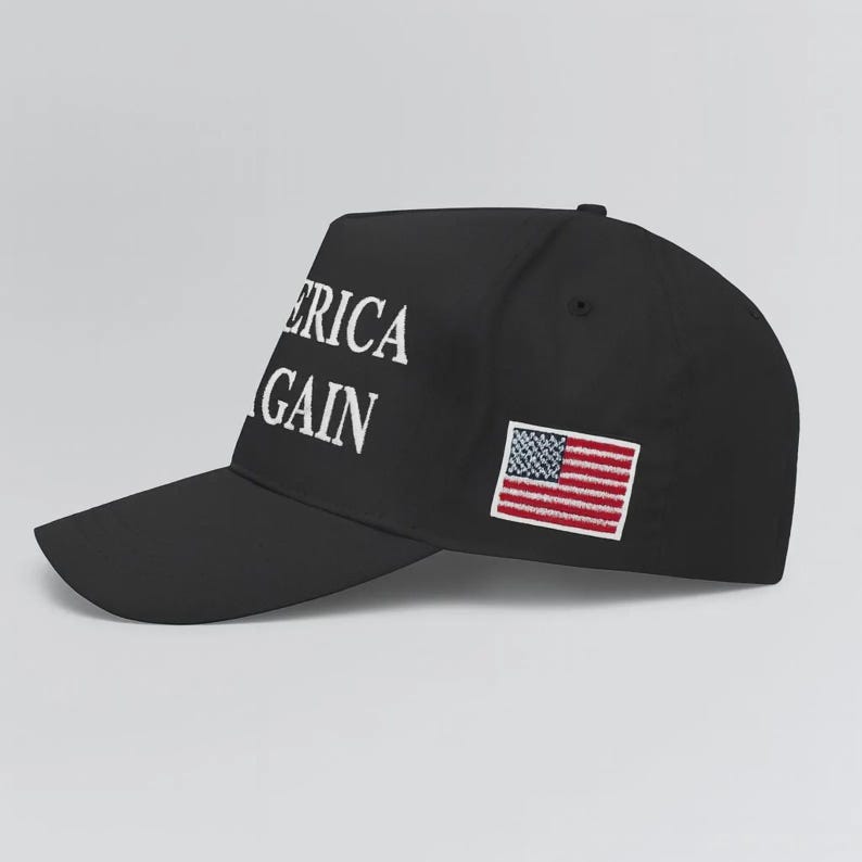 Op de afbeelding: Een zwarte honkbalpet met witte geborduurde tekst die AMERICA AGAIN leest op het zijpaneel. De pet bevat ook een kleine, rechthoekige geborduurde patch van de vlag van de Verenigde Staten, geplaatst naar de achterkant van het zijprofiel.