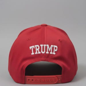 May include: A bright red baseball cap featuring the word TRUMP embroidered in white capital letters on the back panel. The cap includes a matching red plastic snapback closure at the rear and ventilation eyelets on the upper crown panels.
