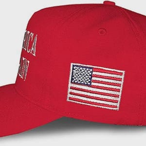 May include: A bright red baseball cap featuring an embroidered American flag patch on the side. The front panel displays partial white embroidered text that reads AMERICA AGAIN. The cap has a structured crown, a curved brim, and a button on top.
