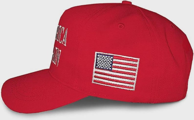 May include: A bright red baseball cap featuring an embroidered American flag patch on the side. The front panel displays partial white embroidered text that reads AMERICA AGAIN. The cap has a structured crown, a curved brim, and a button on top.