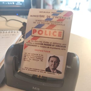 Puede incluir: Una tarjeta de identificación de un inspector de policía francés, con la palabra "POLICE" en rojo y azul. La tarjeta incluye una foto del inspector y está en una base de carga gris. El texto de la tarjeta está en francés.