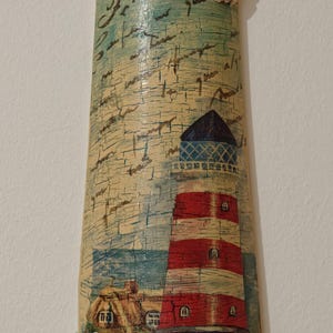Puede incluir: Un adorno de pared rectangular con un faro de rayas rojas y blancas, un tejado azul y una pequeña casa. El fondo tiene un aspecto texturizado y envejecido con conchas y escritura.