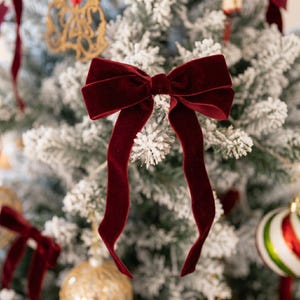 Pode incluir: Um laço de veludo em tom bordô profundo com duas fitas longas e fluidas. O tecido possui uma textura macia e mate que capta a luz, criando reflexos subtis ao longo das dobras e bordas do laço. O design é simétrico com um nó central.