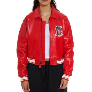 Op de afbeelding: Een rode leren jas met een kraag en ritssluiting. De jas heeft wit en rood gestreepte manchetten en zoom. Een geborduurde patch met de tekst "AVIREX" en een Amerikaans vlagontwerp staat op de borst.