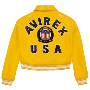 Op de afbeelding: Een felgele bomberjack met het woord "AVIREX" in zwarte letters op de rug. Daaronder een cirkelvormig embleem met "ALL AMERICANS" en "ESTABLISHED 1975". Het woord "USA" staat eronder. De jas heeft wit en geel gestreepte manchetten.