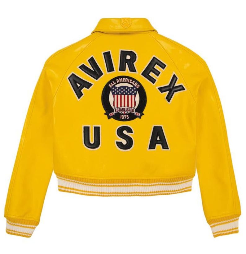 Op de afbeelding: Een felgele bomberjack met het woord "AVIREX" in zwarte letters op de rug. Daaronder een cirkelvormig embleem met "ALL AMERICANS" en "ESTABLISHED 1975". Het woord "USA" staat eronder. De jas heeft wit en geel gestreepte manchetten.