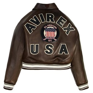 Op de afbeelding: Bruine leren jas met het woord "AVIREX" en "USA" op de rug. De jas heeft een patch met de Amerikaanse vlag en de woorden "ALL AMERICANS ESTABLISHED 1975". De manchetten zijn wit en bruin gestreept.
