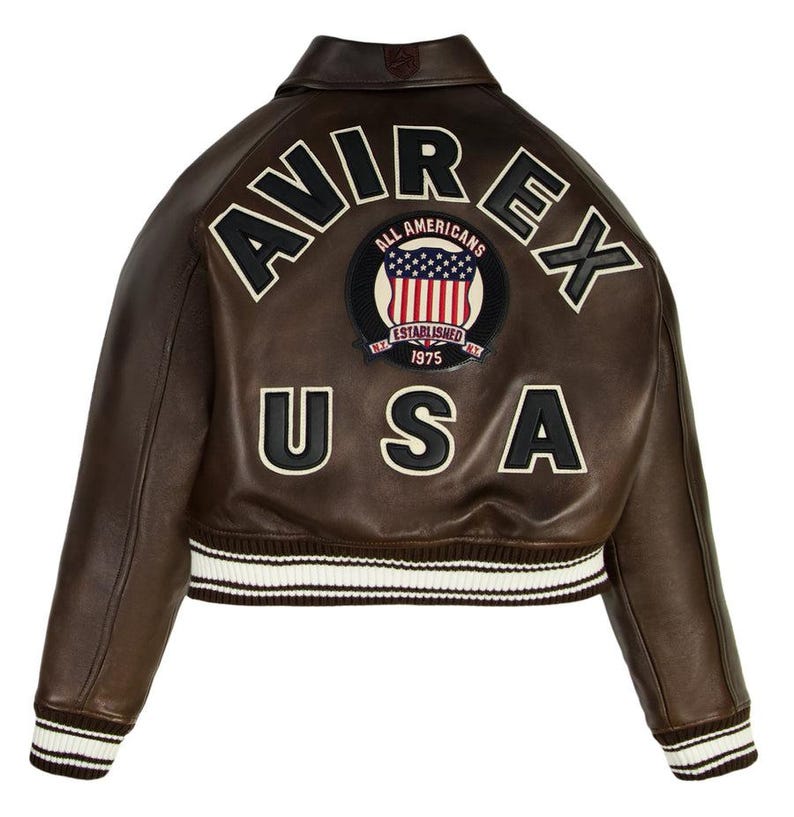 Op de afbeelding: Bruine leren jas met het woord "AVIREX" en "USA" op de rug. De jas heeft een patch met de Amerikaanse vlag en de woorden "ALL AMERICANS ESTABLISHED 1975". De manchetten zijn wit en bruin gestreept.