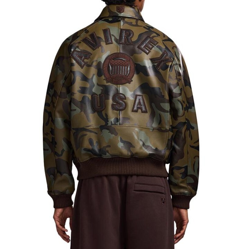 May include: A camouflage bomber jacket with brown detailing. The back displays "AVIREX USA" in brown lettering, with a brown emblem. The jacket has a ribbed hem and cuffs. The jacket is a stylish outerwear piece.