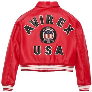 Op de afbeelding: Rode leren jas met het woord "AVIREX" in zwart-witte letters op de rug. Daaronder een patch met "ALL AMERICANS ESTABLISHED 1975" en "USA". De jas heeft een wit en rood gestreepte zoom.