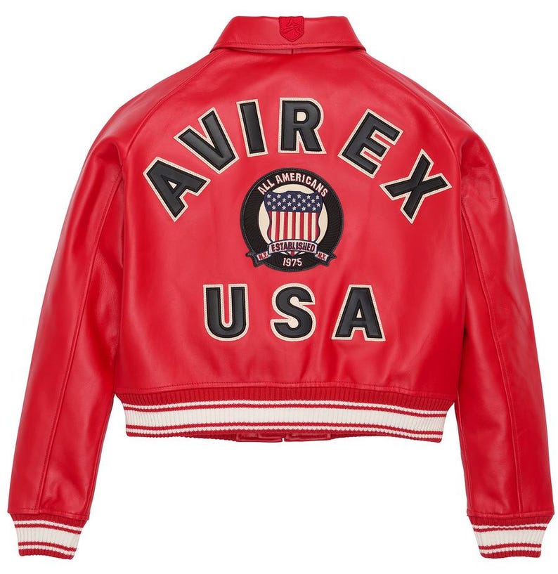 Op de afbeelding: Rode leren jas met het woord "AVIREX" in zwart-witte letters op de rug. Daaronder een patch met "ALL AMERICANS ESTABLISHED 1975" en "USA". De jas heeft een wit en rood gestreepte zoom.