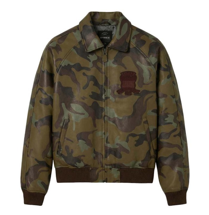 May include: A camouflage leather jacket in green, brown, and tan. The jacket has a collared neckline, a zip closure, and a brown ribbed hem and cuffs. A brown patch is sewn on the chest. The brand name "AVIREX" is visible on the collar.