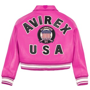 Op de afbeelding: Een felroze bomberjack met witte en zwarte details. De achterkant toont het woord "AVIREX" boven een cirkelvormig embleem met de tekst "ALL AMERICANS" en "ESTABLISHED 1975", en "USA" eronder. Geribbelde manchetten en zoom.