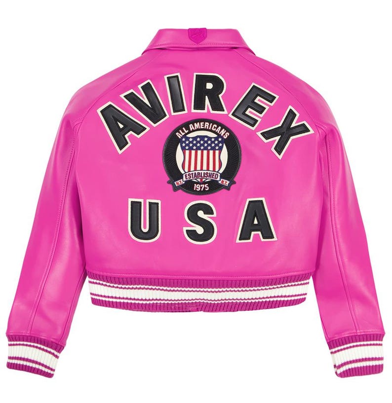 Op de afbeelding: Een felroze bomberjack met witte en zwarte details. De achterkant toont het woord "AVIREX" boven een cirkelvormig embleem met de tekst "ALL AMERICANS" en "ESTABLISHED 1975", en "USA" eronder. Geribbelde manchetten en zoom.