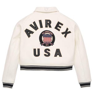 Op de afbeelding: Cr&egrave;mekleurige bomberjack met zwart-witte gestreepte boord. De achterkant heeft de merknaam "AVIREX" in zwarte letters boven een cirkelvormige patch met de Amerikaanse vlag en het woord "USA".