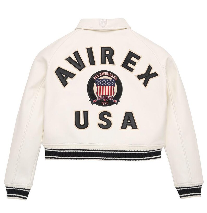 Op de afbeelding: Cr&egrave;mekleurige bomberjack met zwart-witte gestreepte boord. De achterkant heeft de merknaam "AVIREX" in zwarte letters boven een cirkelvormige patch met de Amerikaanse vlag en het woord "USA".