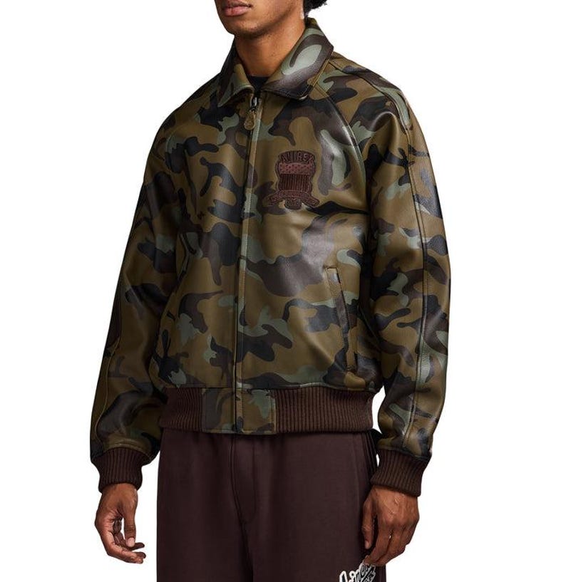 May include: A camouflage jacket in green, brown, and black. The jacket has a collared neckline, a front zip, and brown ribbed hem and cuffs. A patch with text is visible on the chest. The jacket appears to be made of a leather-like material.