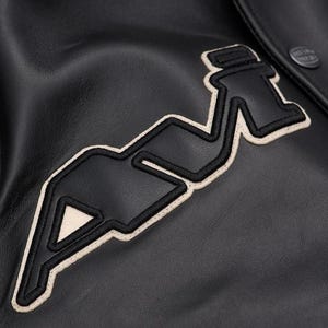 May include: Close-up of a black leather jacket with a large, stylized "A" emblem. The emblem is outlined in off-white stitching and filled with black. A button is visible on the upper right. The jacket has a smooth, textured surface.