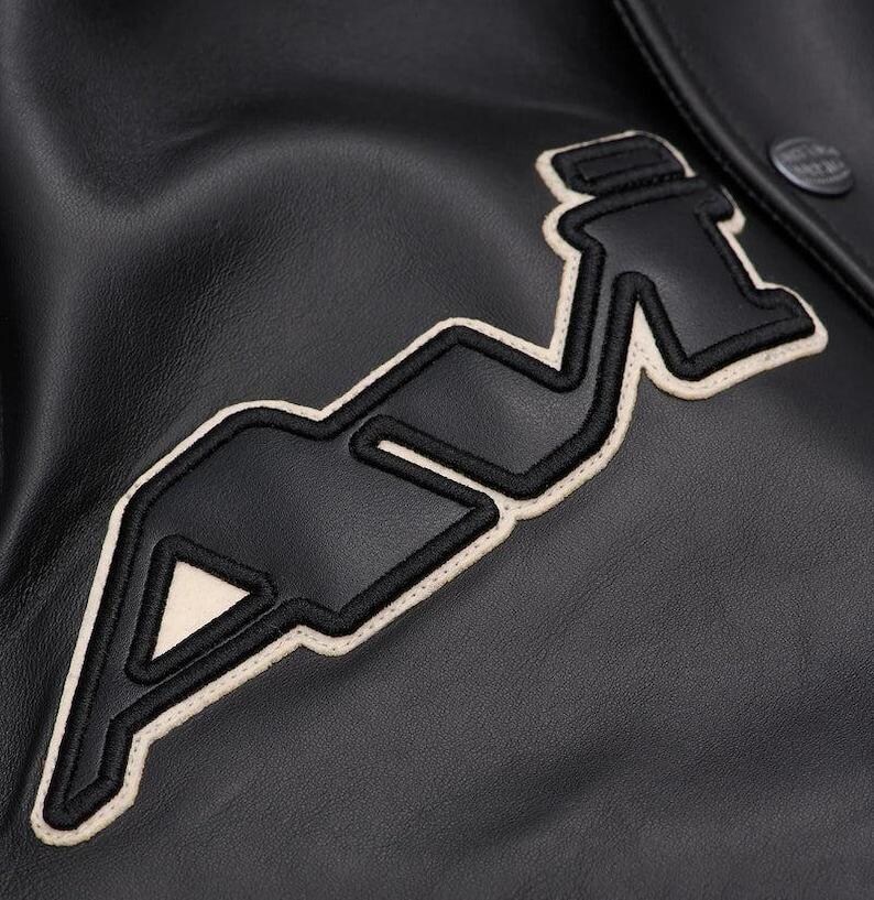 May include: Close-up of a black leather jacket with a large, stylized "A" emblem. The emblem is outlined in off-white stitching and filled with black. A button is visible on the upper right. The jacket has a smooth, textured surface.