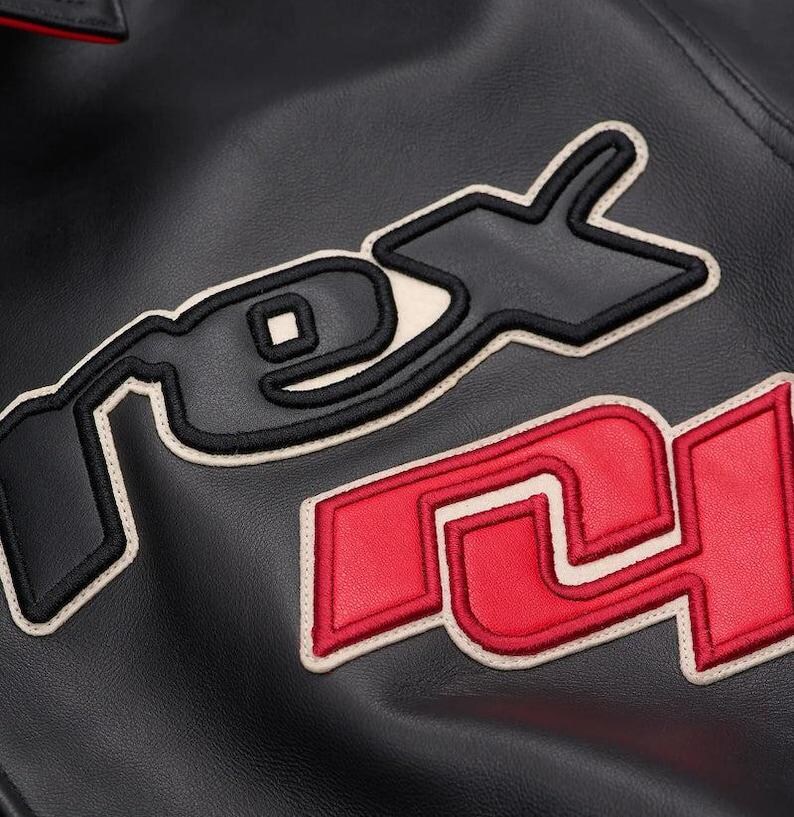 May include: Close-up of a black leather jacket with embroidered patches. The patches feature the letters "RS" in red and "PEX" in black, outlined in cream. The jacket has a smooth texture and a subtle sheen.