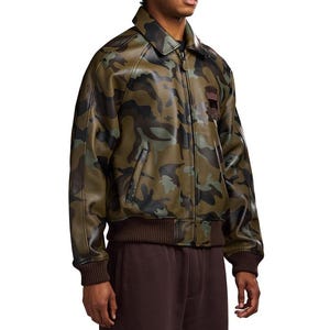 May include: A camouflage-patterned jacket in shades of green, brown, and black. The jacket has a collared neckline, a zip closure, and ribbed hem and cuffs. A small patch is visible on the upper chest. The jacket is made of a leather-like material.