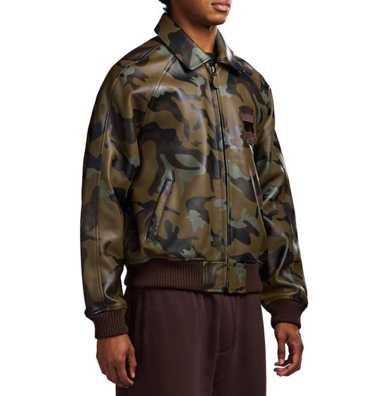 May include: A camouflage-patterned jacket in shades of green, brown, and black. The jacket has a collared neckline, a zip closure, and ribbed hem and cuffs. A small patch is visible on the upper chest. The jacket is made of a leather-like material.