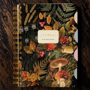 May include: A spiral-bound journal with a black cover featuring a botanical illustration of mushrooms, ferns, and berries. A central cream-colored label displays the text JOURNAL and BV by Bruno Visconti. The notebook includes colored side tabs.