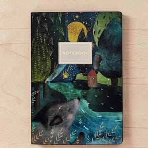 May include: A rectangular notebook with a dark, illustrated cover featuring a wolf looking up at a large yellow moon in a starry night sky. The scene includes green trees, a small house, and a river. A central rectangular label reads NOTEBOOK.