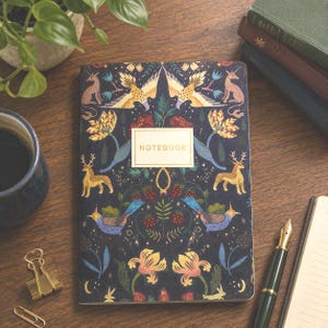 May include: A rectangular notebook with a dark blue cover featuring a symmetrical pattern of golden deer, birds, mythical creatures, flowers, and strawberries. A central rectangular label with a gold border contains the word NOTEBOOK in gold.
