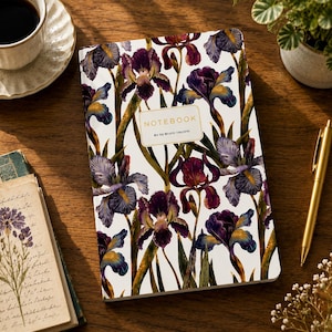 Può includere: Un taccuino rettangolare con copertina bianca caratterizzato da un motivo ripetuto di fiori di iris viola e gialli. Un'etichetta rettangolare centrale sulla copertina riporta il testo NOTEBOOK e By by Bruno Visconti in lettere dorate.