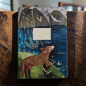May include: A rectangular notebook featuring a printed illustration of a brown spotted animal standing on a green grassy bank near a dark blue body of water. In the background, large gray rock formations and white birds are visible. The word NOTEBOOK is printed in gold text within a central rectangular frame.