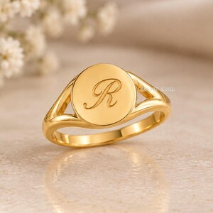 Op de afbeelding: Een gouden zegelring met een cirkelvormig vlak waarin de letter R in cursief schrift is gegraveerd. De ring heeft een gepolijste afwerking en een gespleten schachtontwerp dat naar de achterkant van de band toe taps toeloopt.