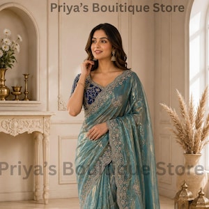 Op de afbeelding: Een blauwgroene sari met een metallic glans en ingewikkeld zilveren borduurwerk langs de randen, gecombineerd met een donkerblauwe blouse versierd met gedetailleerde zilveren bloemmotieven. De tekst Priya’s Bouitique Store is zichtbaar.