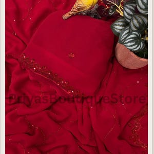 May include: A vibrant red fabric featuring a delicate pattern of thin, shimmering lines and small sequins. The material is draped with soft folds and includes a decorative border with intricate red beadwork. The text PriyasBoutiqueStore is visible.