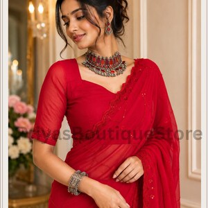 May include: A vibrant red saree featuring a sheer fabric with delicate tonal embroidery and sequin accents. The ensemble includes a matching red blouse with short sleeves, a statement silver-toned choker necklace with red inlays, and a wide silver cuff.